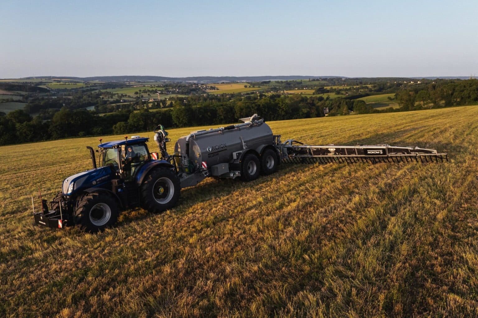 Aerial view of a tractor pulling a Pichon SC20R slurry tanker with applicator extended across a hillside field